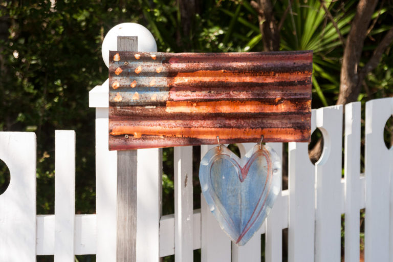 A US flag made from corrugated metal on a wooden pole, with a heart shaped drawing attached to. It sits next to a fence in front of a house