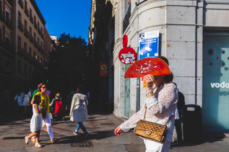 A wopman with a red fan walking on the street. She has a gold handbag. Other people are walking around in the background.