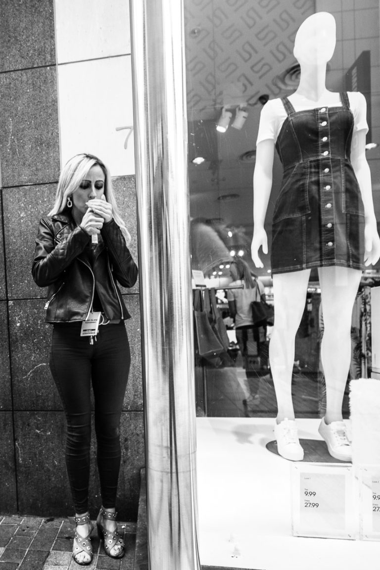 A woman standing in the street lighting a cigarette next to a window with a mannequin