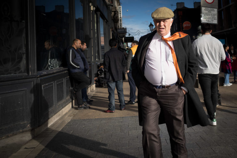 A man walking on the street. The breeze blows his orange tie over his shoulder. People are visible in the background.