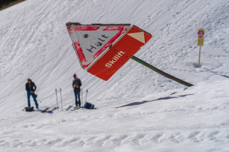 Two skiers prepare to go downhill, while in the foreground a sign warns "Halt" and "Skilift" pointing to the right but the sign is leaning precariously to the side