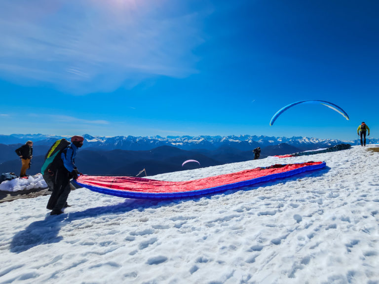A paraglider adjusts their wing on the ground on top of a mountain in Germany. A blue sky above and mountains in the distance.