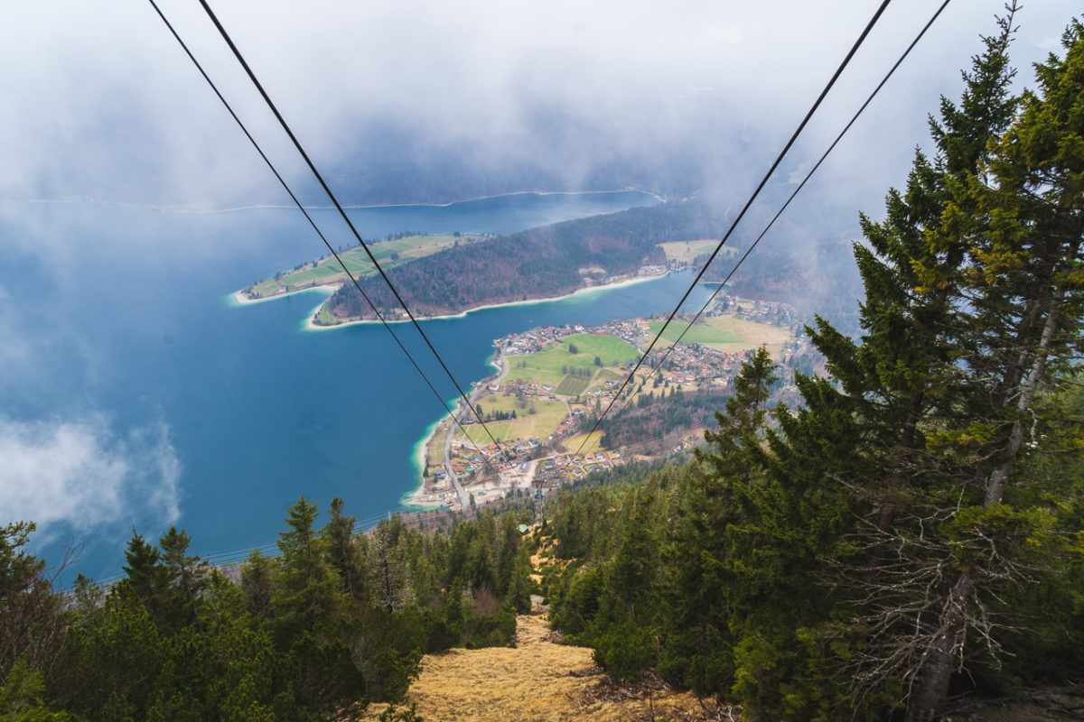 The view from the gondola station high up over a valley. Blue water of a lake is seen below as well as tiny houses, roads and trees surrounding green fields.