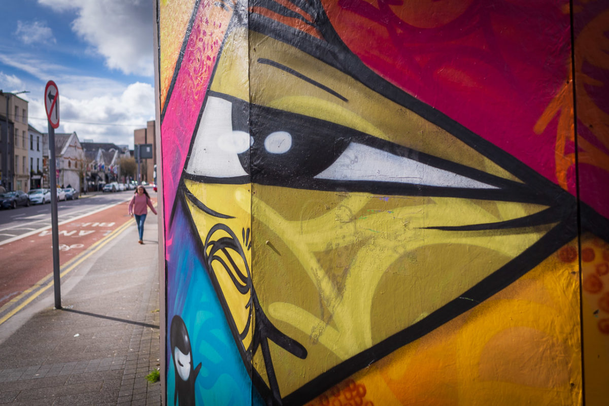 Hoarding around a derelict site in Cork City has graffiti on it and an eye looks out of the corner while a young woman walks up the street