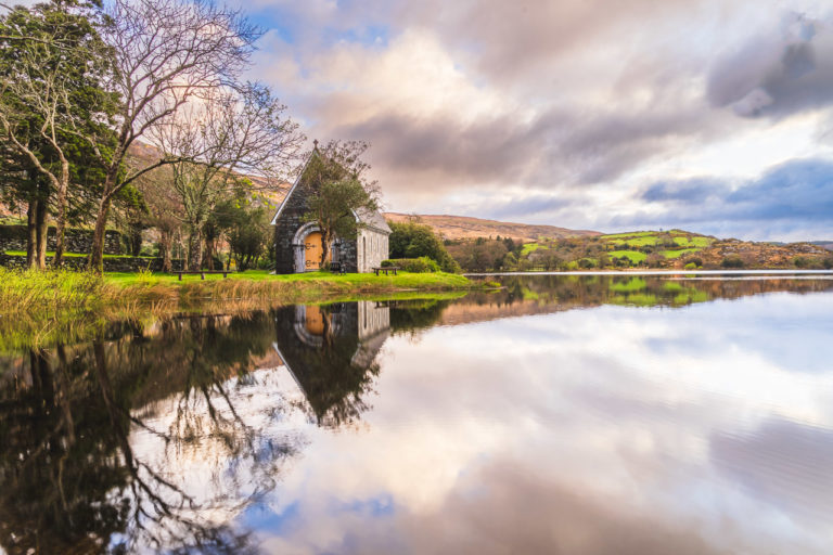 The small chapel at Gougane Barra reflected in the lake there in early morning light. Trees are near it and fields and hills in the distance. It's a cloudy day.