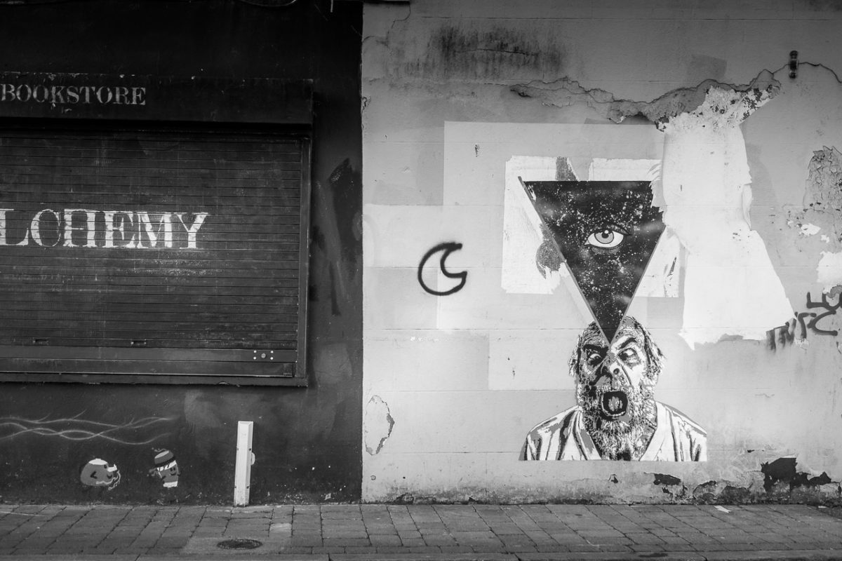 Graffiti on a wall. An eye in a triangle with a starry background overlaid on the face of a screaming bearded man.
