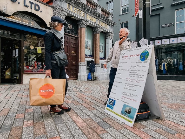 A man on the street with a large sign warning "Global Warning". A woman carrying a shopping bag is listening patiently.
