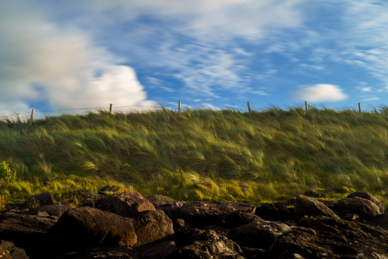 Clouds blur with movement, and grass bends against the wind in a long exposure photo at a beach in Kerry. Rocks in the foreground, Grass on a hill, with a broken fence on top, and the cloudy sky above.