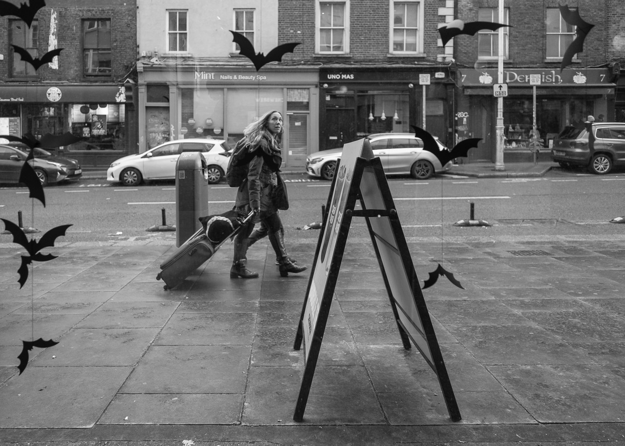 A woman passes, looking in at the Bubble Tea shop I'm looking out of. Plastic bats are flying around, suspended on thing string, to celebrate Halloween.