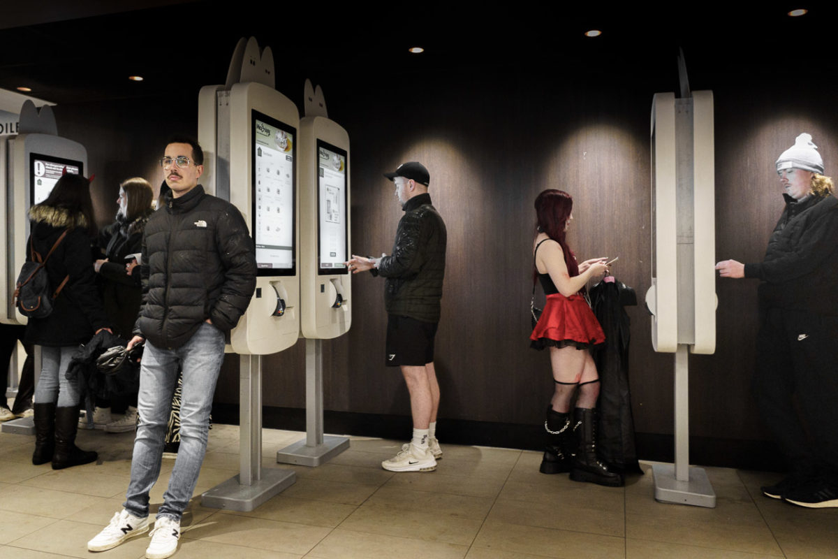 People ordering food in McDonalds using their kiosks. Four people are at the machines, mostly dressed for the night in dark clothes and coats while one young woman is dressed as if it wasn't cold and wet outside.