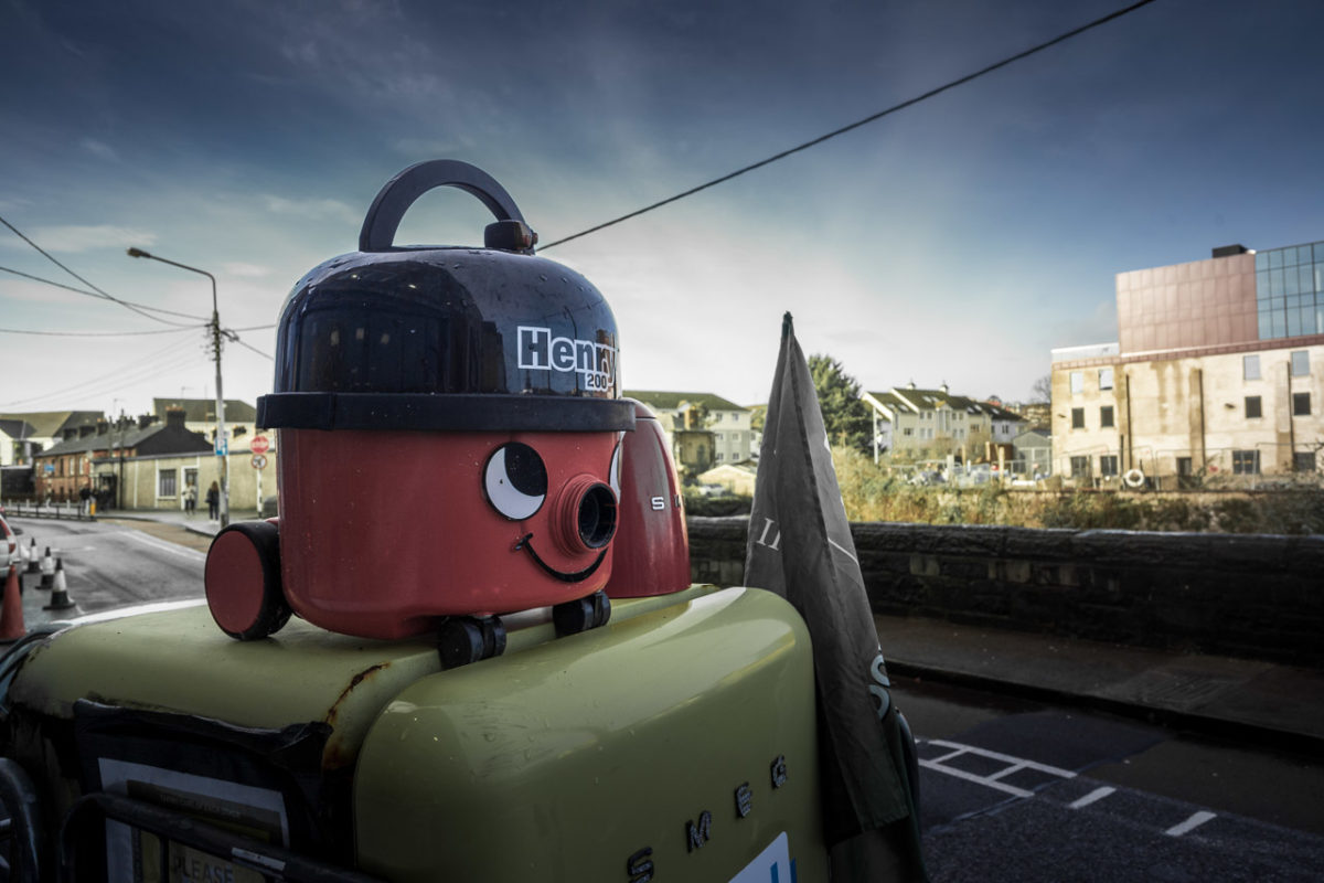 A Henry vacuum cleaner on top of an old style fridge out on the street in front of a hardware store