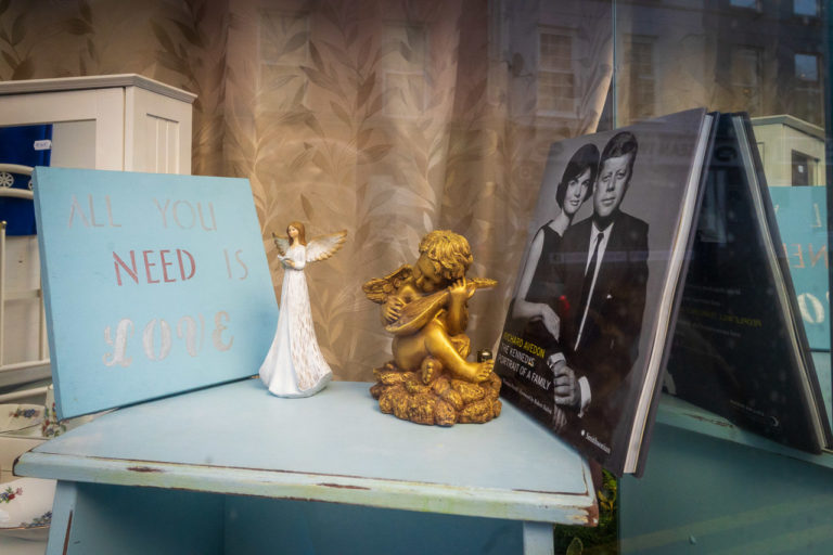 A shop front: a table with a sign that says "All you need is love", a statue of an ange, a cherub statue and the book, "The Kennedys Portrait of a family" by Richard Avedon.