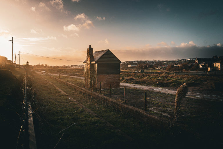 The old railway line to Youghal in the setting sun. A small house in the middle distance and poles and cables off further and houses off in the background.