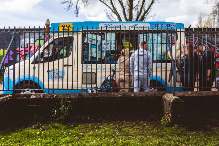 Parents and I presume children (unseen), line up next to an ice cream van outside a park with railings.