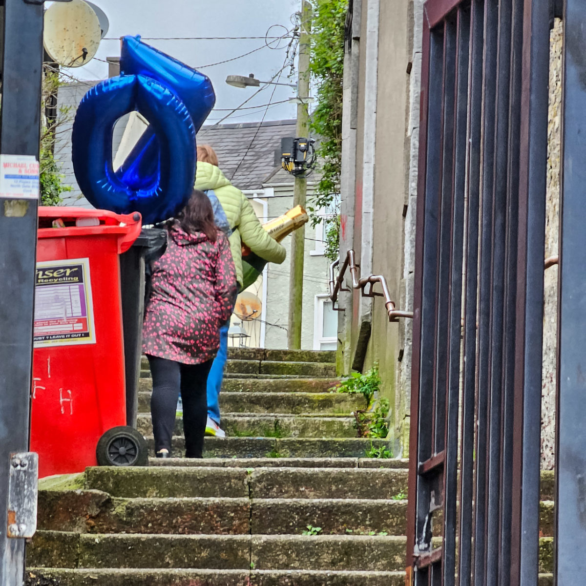 Two people walking up the steps of a narrow street. They have two balloons in the shape of a 7 and a 0, and a large fake bottle of champagne.