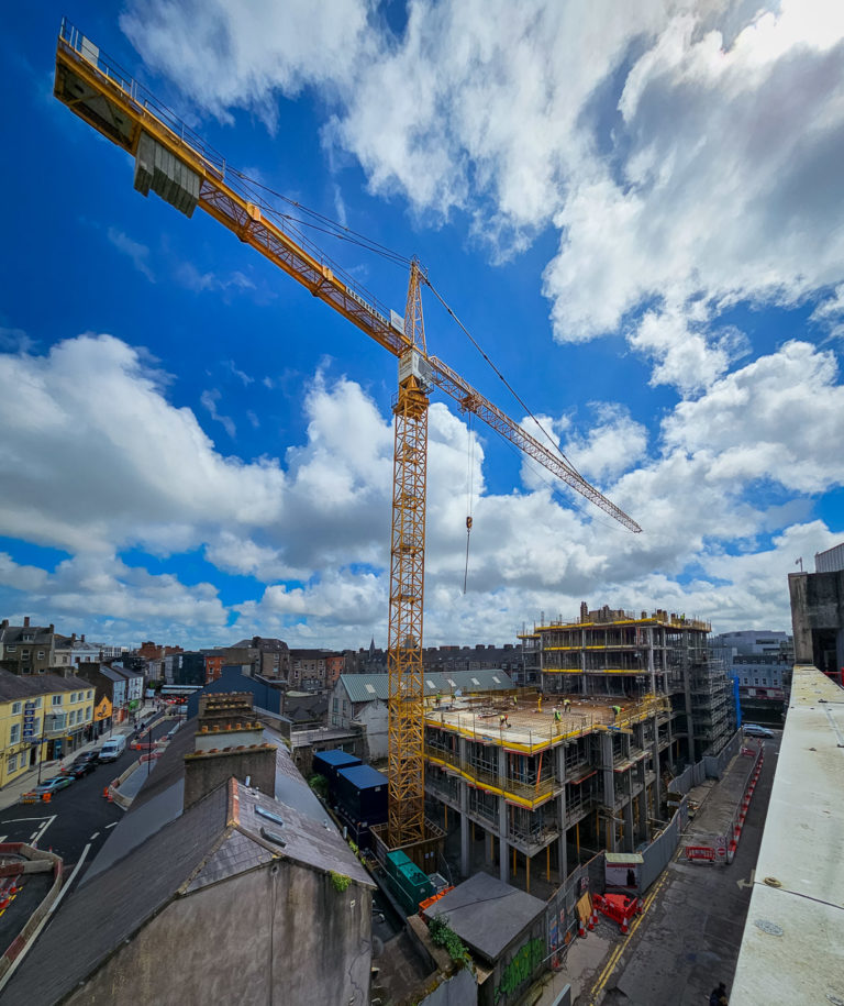 Construction work on a building site in Cork City. A tall yellow crane is visible under a sky half full of clouds. Tiny figures of men can be seen on the building site working on laying down steel supports.
