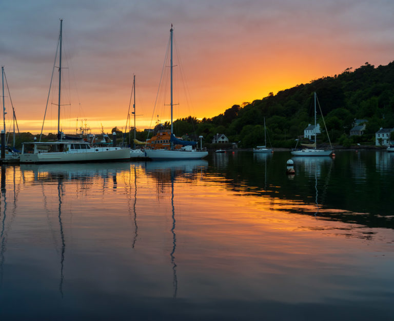 Boats on the water and in the background a forest with some houses and a lovely orange sunset too.