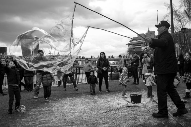 A man created huge bubbles in London with two poles and string with little loops, and one huge loop. Adults and kids watch and try to burst them.