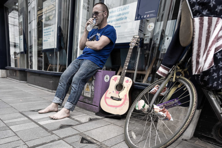 A barefoot man takes a break from playing the guitar on the street. He is drinking some water while his guitar sits next to him and a bike leans against the wall.