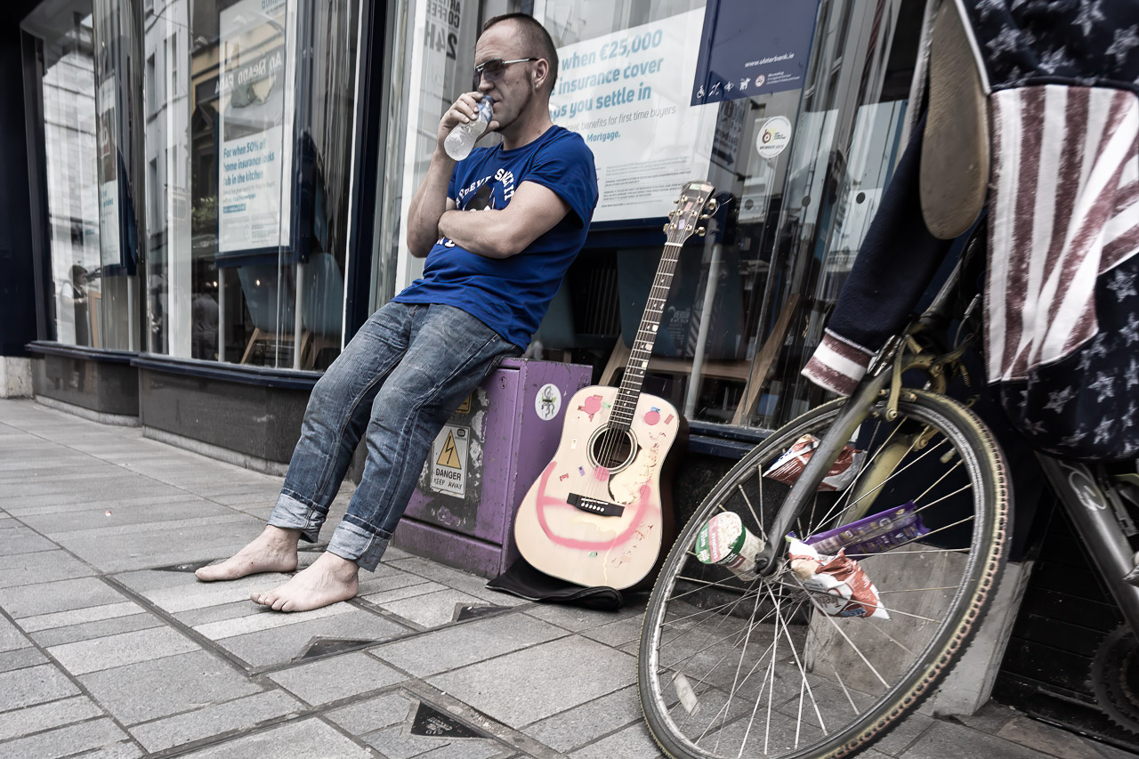 A barefoot man takes a break from playing the guitar on the street. He is drinking some water while his guitar sits next to him and a bike leans against the wall.