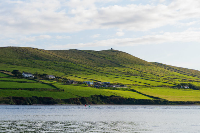 The sun shines on the headland opposite Dingle in Co Kerry. Clouds cast a shadow over the fields on a glorious sunny day. The water of Dingle Bay is visible in the foreground.