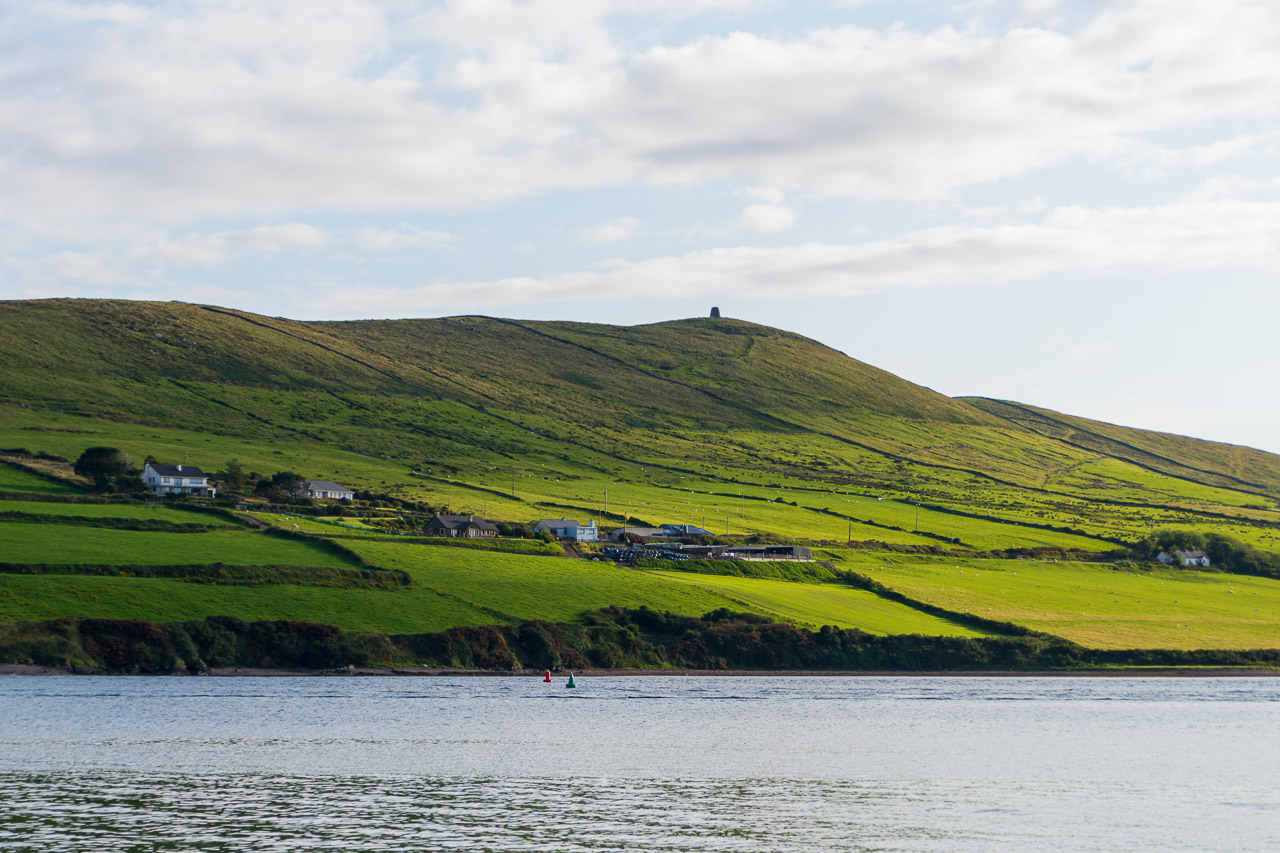 The sun shines on the headland opposite Dingle in Co Kerry. Clouds cast a shadow over the fields on a glorious sunny day. The water of Dingle Bay is visible in the foreground.