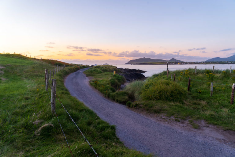 A road curves off into the distance towards some parked vehicles on a headland. Water is visible to the side and the sky is turning orange at the horizon as the sun has set