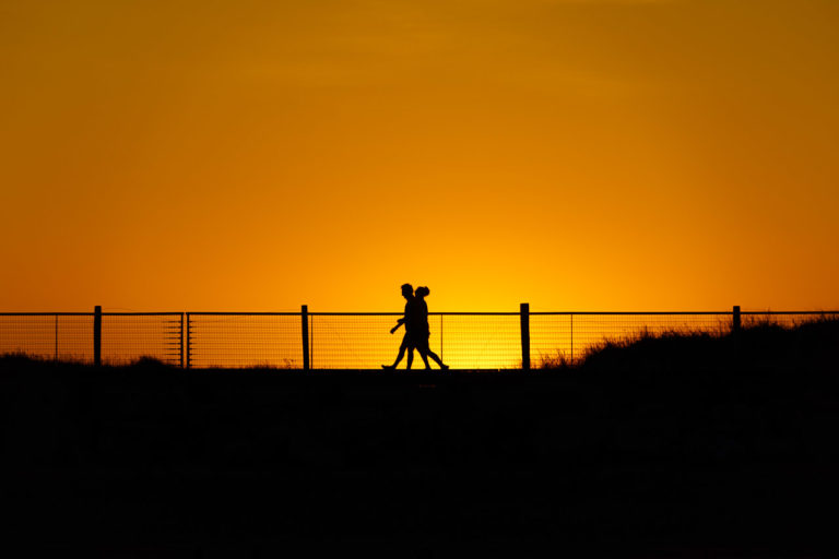 Silhouettes of two people walking on the Youghal boardwalk, with the sun setting behind them