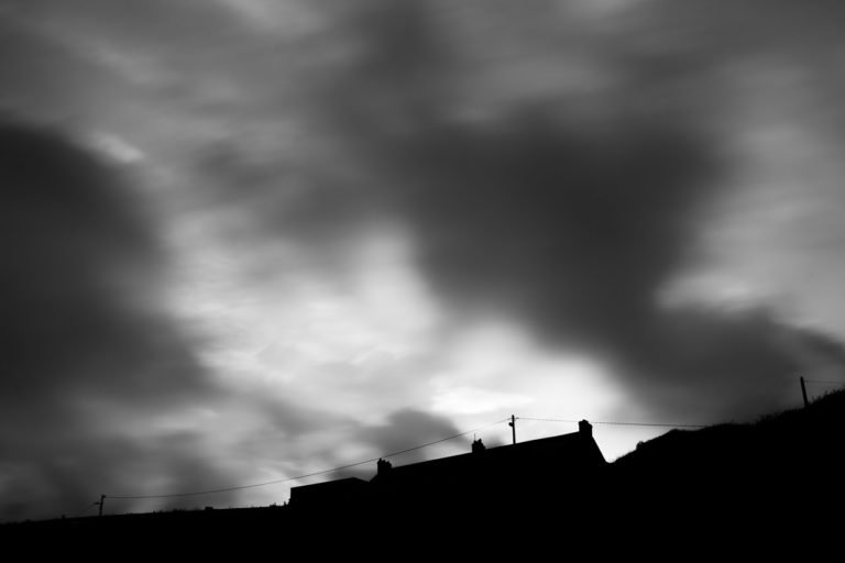 Wind blows the clouds over a silhouetted house on a hill in a black and white photo.