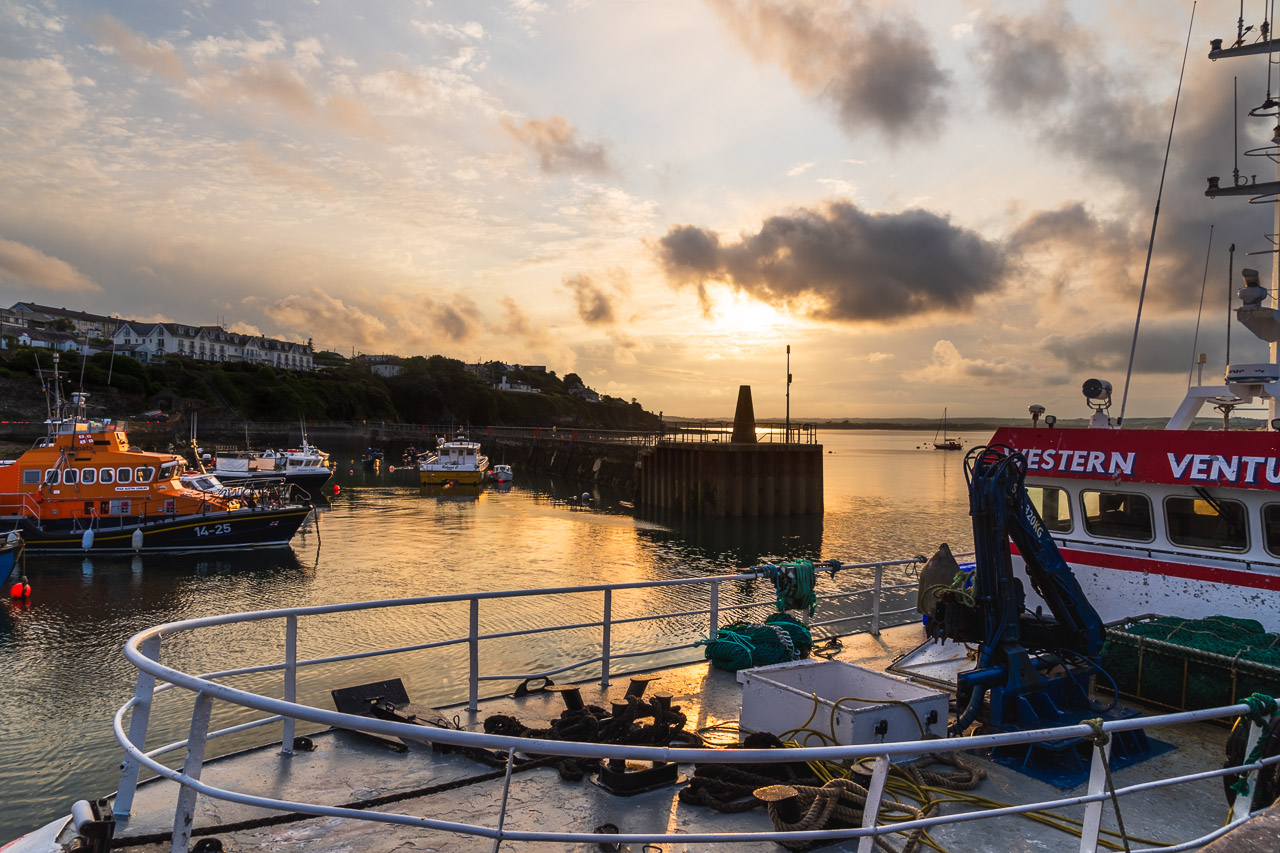Boats in Ballycotton Harbour at sunset. The lifeguard vessel can be seen in the middle background, and a trawler is tied to the pier right in front of me, with other smaller boats further back.