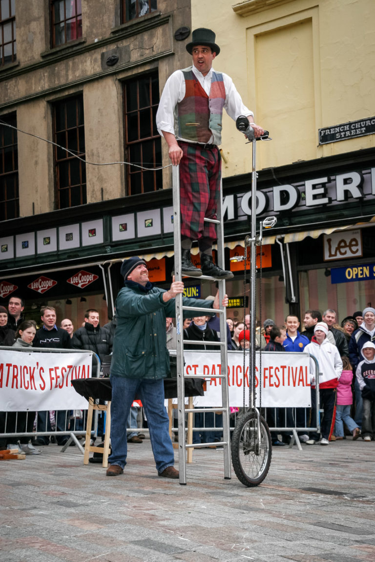 A man gets on a very tall unicycle with the help of a ladder and a passer by, watched by a crowd all around him.
