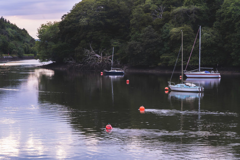 3 boats tied up on the river between Drakes Point and Crosshaven. Buoys are visible in the water and the opposite bank is covered in trees.