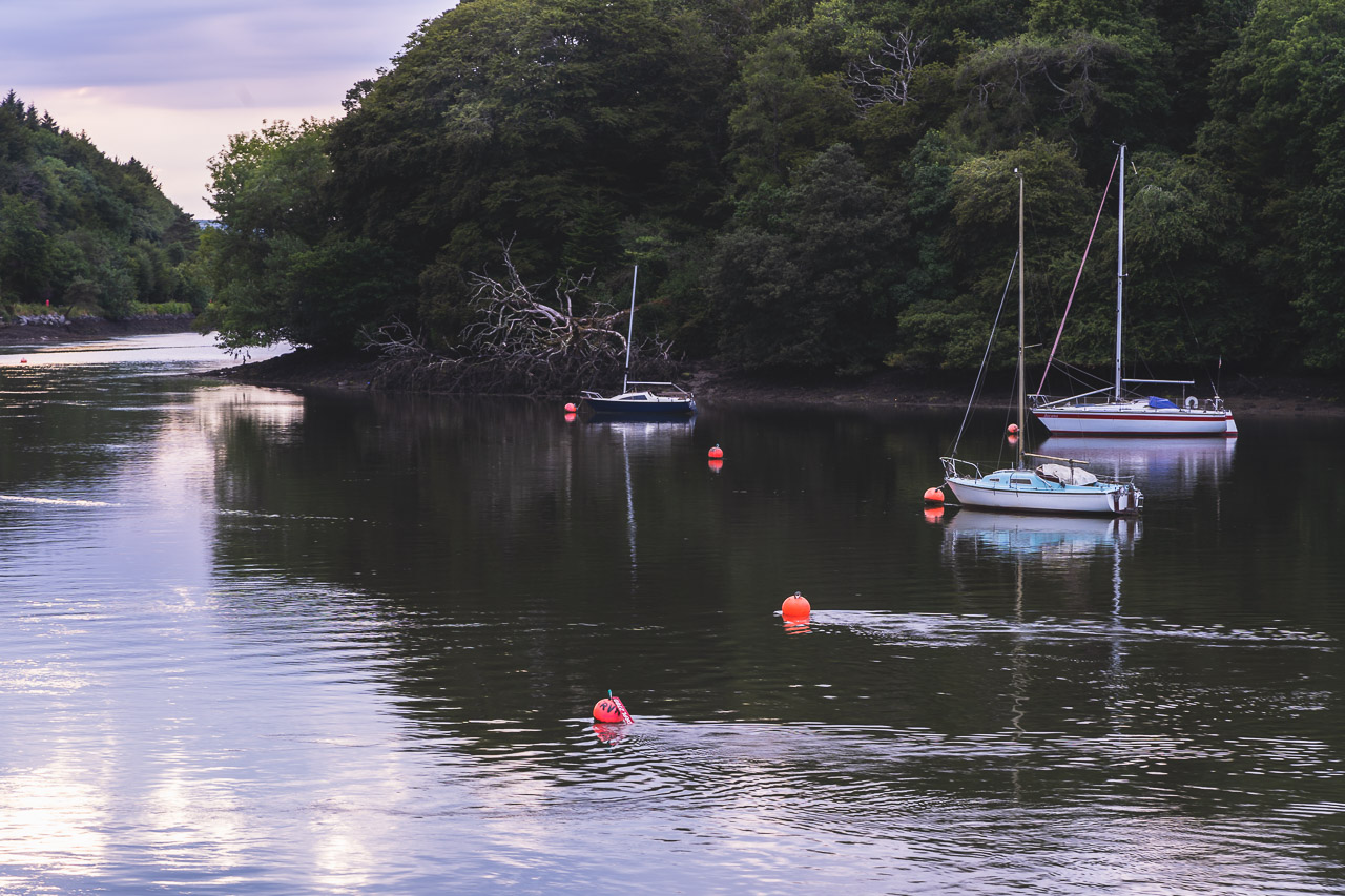 3 boats tied up on the river between Drakes Point and Crosshaven. Buoys are visible in the water and the opposite bank is covered in trees.