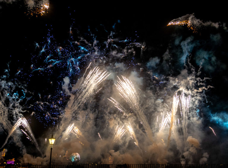 Fireworks light up the sky. A railing in the foreground connects to an ornate lamp post and light and a globe is visible in the background.