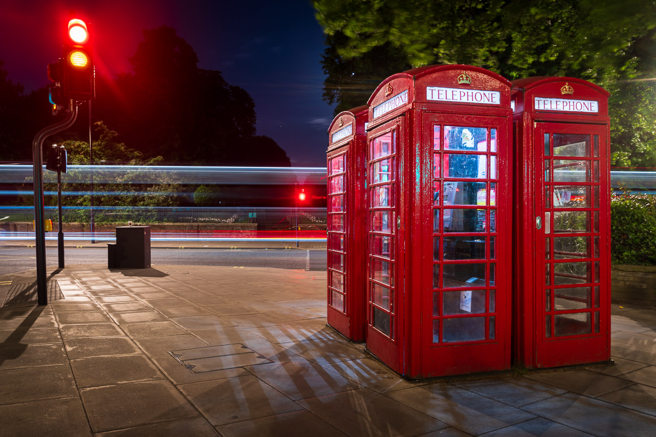 A long exposure photo at night. Light trails from passing vehicles in the background while in front are 3 phone boxes and traffic lights showing red and amber lights.