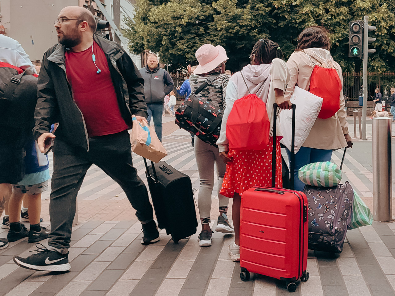 A man dragging a small suitcase hurries off to the left at a pedestrian crossing. He's wearing a red tshirt. A family with suitcases and bags are about to cross. One of their suitcases is red, and two of their bags are. A teenage girl in the group is wearing a red spotted dress.