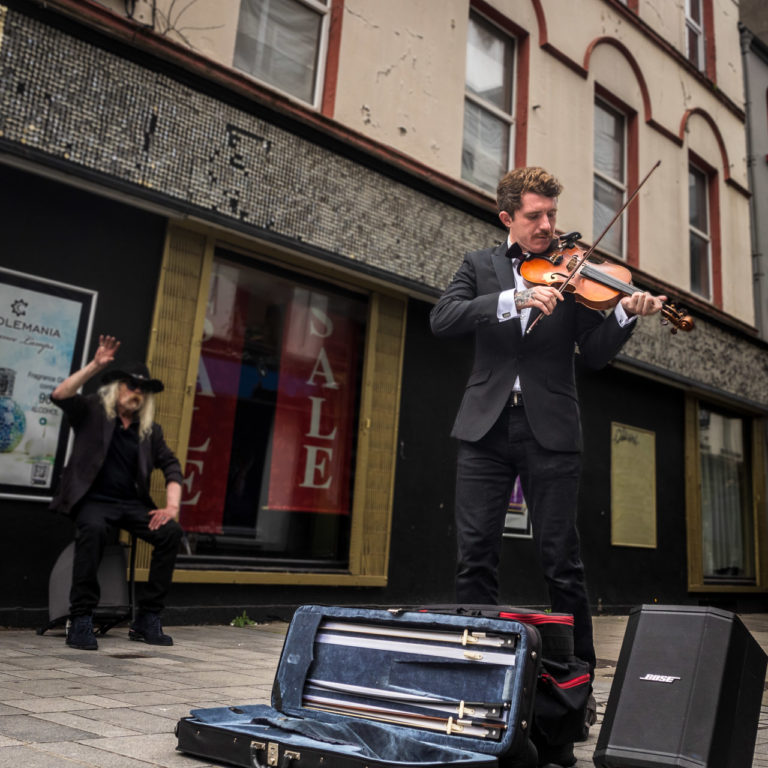 A man playing a violin on the street, with an older man sitting in the background with his arm raised, enjoying the music