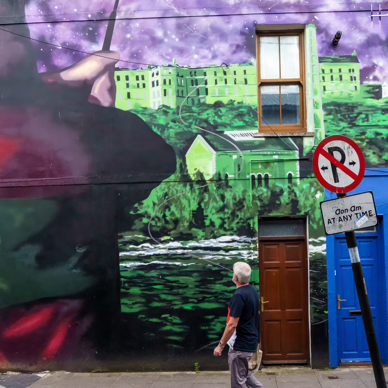 A man looks up at a large mural on a wall. Only part of it is visible but it's a person fishing on a river, with woods and buildings on the far bank.