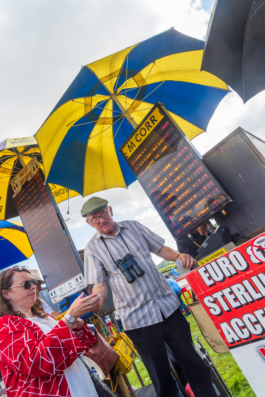A woman takes a receipt after placing a bet at a horse race. The bookie stands on a platform, with a screen and umbrella protecting him.