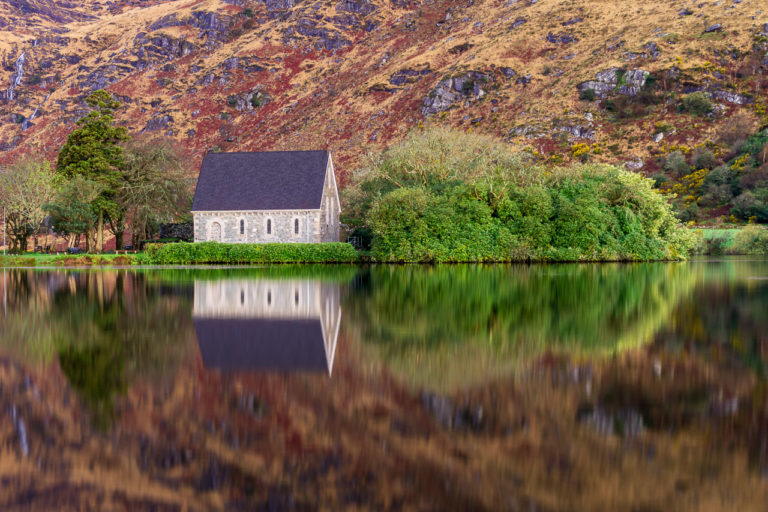 The chapel on the island at Gougane Barra is reflected in the waters of the lake there. Bushes and trees surround it and a mountain takes up the background