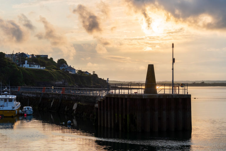 The breakwater protecting boats in Ballycotton harbour lies in shadow as the setting sun shines down behind it.