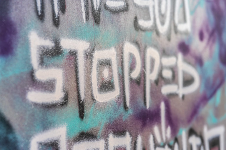 Graffiti on the hoarding around a derelict site says "STOPPED". The tops and bottoms of other characters are visible in the top and bottom of the photo. The background is a mixure of blues, purples and grey