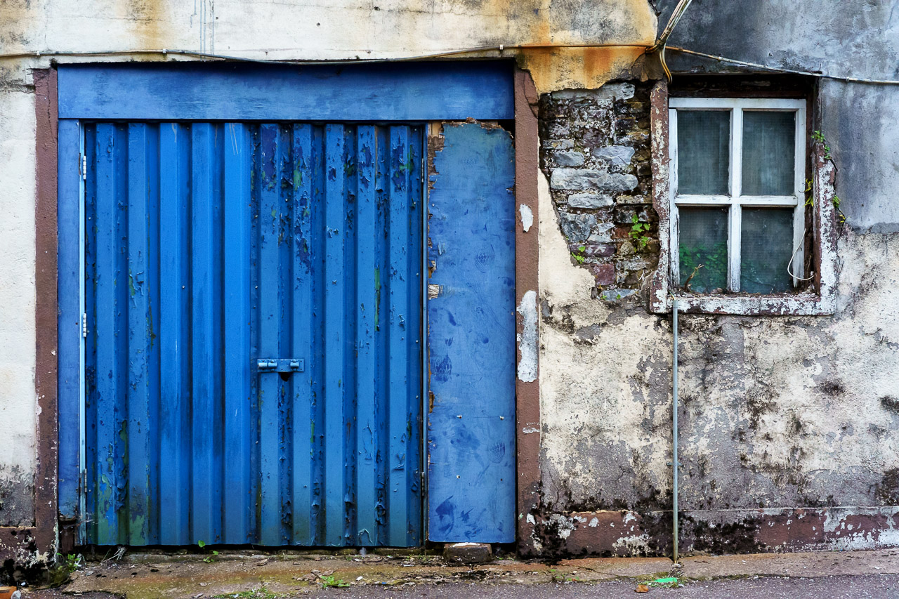 A blue door in a dilapidated building in need of repair. Masonry is crumbling and a nearby window has seen better days.