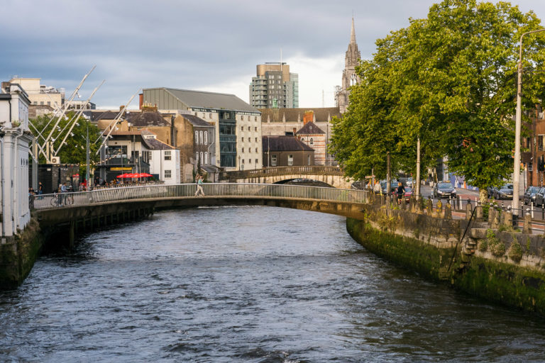 A footbridge called Nano Nagle Bridge crosses the River Lee. The river runs underneath in this photo. Trees are visible on the right. Buildings all around.