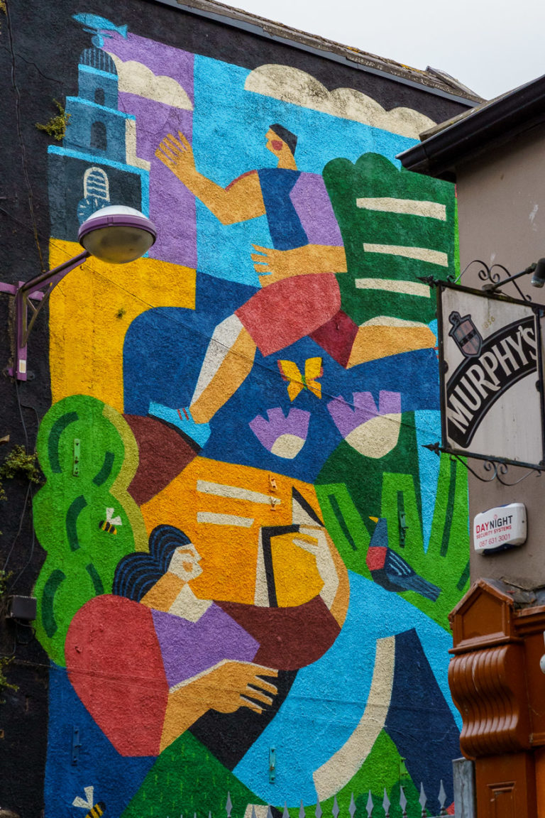 A brightly coloured mural showing a man running, a woman reading and Shandon Bells. A light is visible and a pub sign for Murphy's Stout