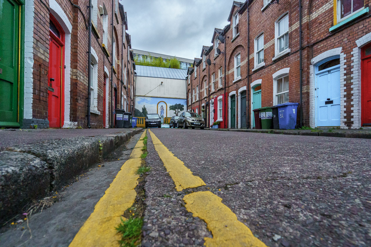 A photo of a street from low down by the ground. The yellow lines next to the pavement lead off to the end of the street. Cars are parked on the road and houses are old, red brick buildings. A (Catholic?) mural featuring Mary is painted on the wall at the end of the street.