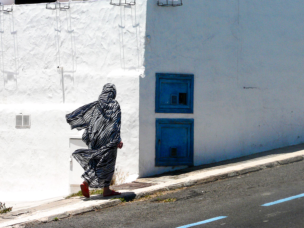 A woman walks up a hill in Lanzarote. She's cover head to ankles in flowing cloth with black and white stripes. A white washed house is behind and she's about to walk from the glare of the sun into the shadow of the house.