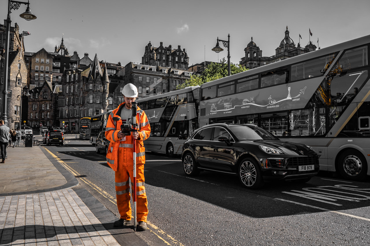 A worker dressed in orange overalls and a hard hat inspects a device he's working on while standing at the edge of a busy road. Cars and busses behind him.
