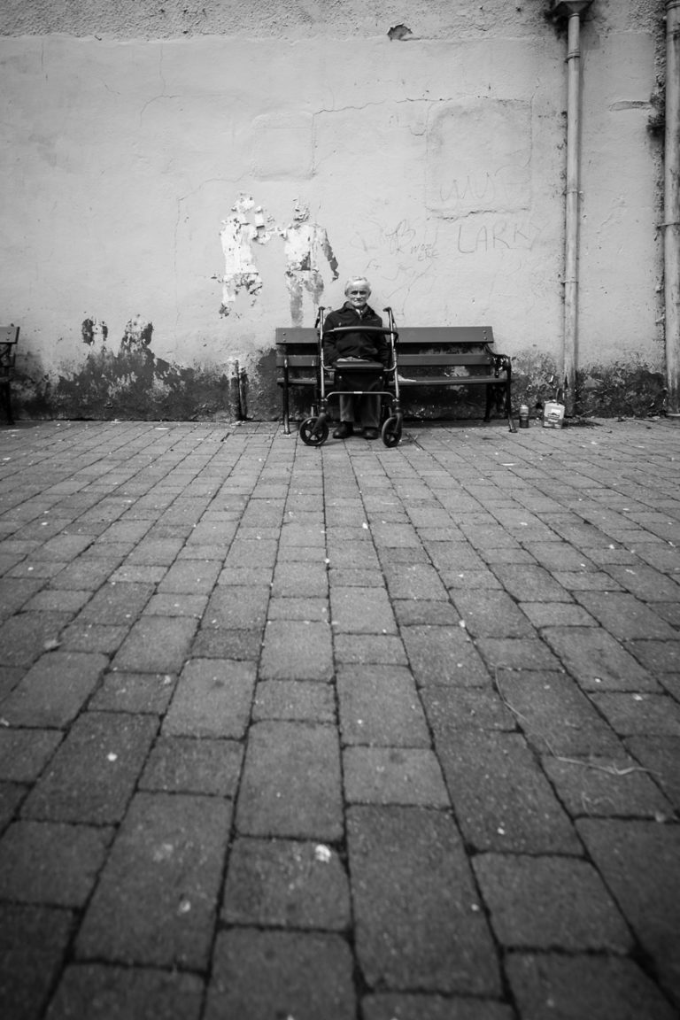 An elderly man sits on a bench in Bishop Lucey Park. The wall behind him is normally a mural but in July 2019, the wall was mostly blank.