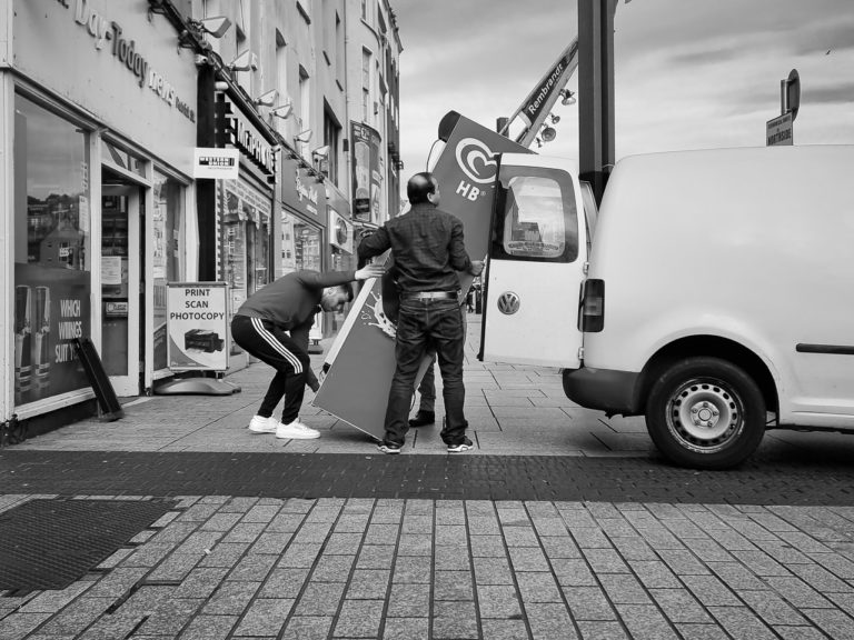 An ice cream fridge is loaded into a van from a shop on Patrick's Street, Cork. Two men hold the mid section while another man bends down to lift it up. The end of a van and it's doors are visible on the right.